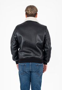 Giubbotto bomber in pelle nera con colletto in shearling bianco, polsini e vita a costine, indossato sopra jeans in denim blu, visto da dietro.