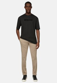 Mustapaita lyhyillä hihoilla, jossa on mustalla painettuna "BOGGI", yhdistettynä beigeihin housuihin ja mustiin lenkkareihin, esittelee rentoa tyyliä.