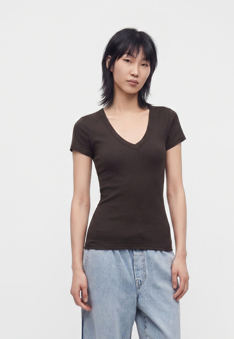 Jeune femme aux cheveux noirs raides portant un t-shirt marron foncé ajusté à col en V et un jean large bleu clair sur un fond uni.