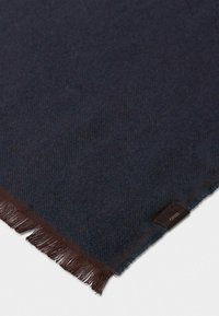 Sciarpa in lana blu navy con motivo a spina di pesce, dotata di bordi frangiati marroni e un piccolo etichetta in tessuto nell'angolo.