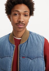 Denim bomber jakna s smešnim rebrastim ovratnikom, s patentnim zatvaračem i crvenim Levi's logotipom na lijevoj strani prsnog koša. Nosena preko crvene majice.