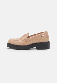 Mocassin en cuir beige avec une semelle épaisse noire, des détails cousus, et une petite étiquette en tissu noir et beige près du talon.