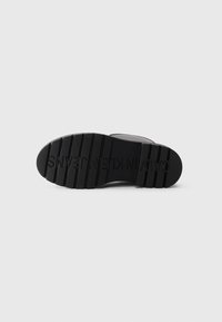 Semelle de chaussure noire avec de profondes rainures et "CALVIN KLEIN JEANS" embossé au bas, présentée sur un fond blanc uni.