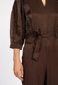 Bruine textuur jumpsuit met een diepe V-hals, driekwartmouwen en een tailleband voor een getailleerde silhoeet. Glad, lichtgewicht materiaal.