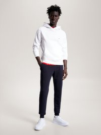 Hoodie branco com bolso frontal, camada interior vermelha, combinado com calças de moletom azul-marinho e sapatilhas brancas, diante de um fundo neutro.