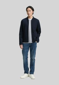 Man in een marineblauwe jas over een wit en marineblauw gestreept shirt, blauwe jeans en witte sneakers, staand tegen een lichte achtergrond.