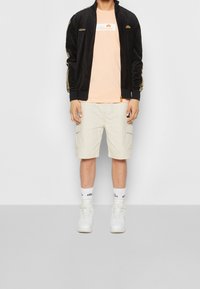 Černá sametová bomber bunda se zlatými akcenty, světle broskvové tričko s logem, béžové cargo šortky, bílé vysoké tenisky a bílé ponožky s logem.