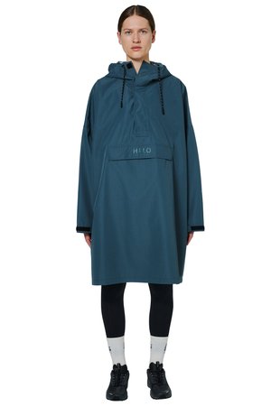 Kvinde iført oversized teal hættetrøje regnjakke med frontlomme, sorte leggings, hvide sokker og sorte sneakers, set forfra.