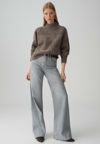 Maglione grigio a maglia testurizzata con collo alto, abbinato a jeans ampi grigi chiari. Una cintura marrone scuro evidenzia il punto vita, taglio classico.