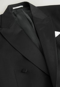 Schwarzer Blazer mit satinierter Oberfläche, Spitzrevers, einem einzigen Knopfverschluss und einem dezent gemusterten Futter. Weißes Taschentuch als Detail.