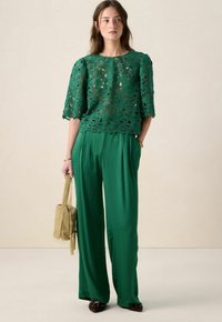 Groen kanten top met bloemmotieven, gecombineerd met losse groene broek. Model houdt een gestructureerde groene handtas vast. Elegant en lichtgewicht ontwerp.