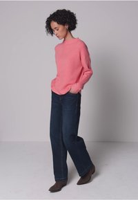 Suéter de punto rosa con un escote amplio, combinado con jeans de pierna ancha en azul oscuro y botines marrones, sobre un fondo gris.