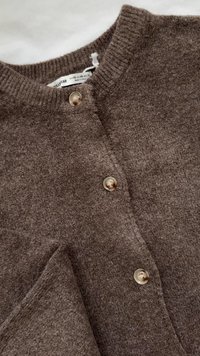 Bruine wollen trui met een geribde halslijn en vijf knopen. De stoftextuur lijkt zacht en pluizig, wat het gebreide ontwerp benadrukt.