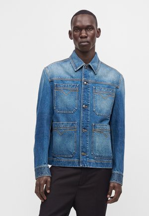 Mand iført en blå denimjakke med fire forlommer og sorte bukser, stående mod en ensfarvet hvid baggrund.