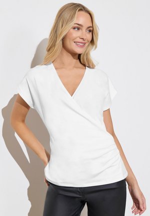 Blonde Frau lächelt, trägt ein weißes V-Ausschnitt-Kurzarmshirt und schwarze Hose, steht mit einer Hand in der Hüfte vor weißem Hintergrund.