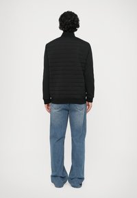 Casaco preto almofadado com um colarinho alto, costuras texturizadas e punhos canelados, combinado com uns jeans de corte largo em azul claro.