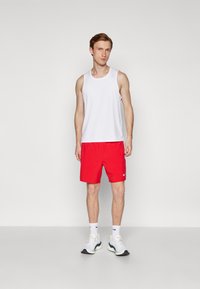 Weißes Tanktop mit kleinem Logo, rote Shorts und weißer Sportschuhe mit Akzenten. Einfaches, sportliches Design, geeignet für aktive Kleidung.