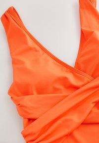 Haut de maillot de bain orange vif style cache-cœur avec bretelles épaisses et détail de tissu froncé sur fond blanc uni.