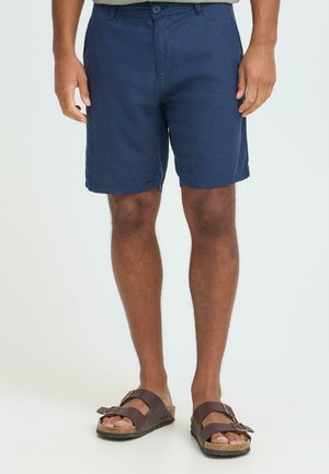 Marineblå shorts laget av et lett stoff, med knappelukking og lommer foran. Brukes med brune sandaler.