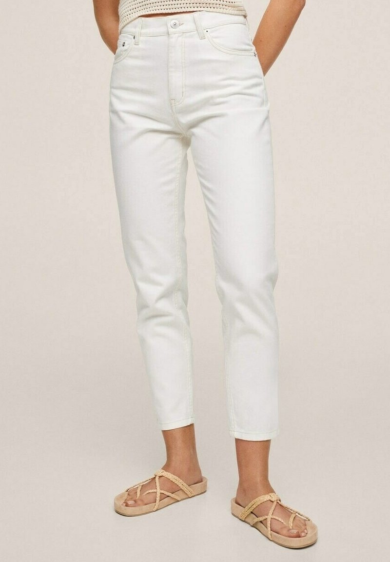Femme portant un pantalon cropped blanc taille haute et des sandales beiges à brides tressées, se tenant devant un fond clair uni.