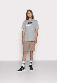 Hallgrey Nike t-särk musta logoga, beežid lühikesed püksid, mustad sneakerid valgete taldadega ja valged sokid mustade triipudega. Ühtlane taust.