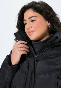 Chaqueta acolchada negra con cuello alto, que cuenta con cierre de cremallera y botones a presión. La tela parece suave con una textura acolchada.