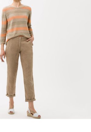 Femme portant un pantalon beige retroussé, un pull rayé beige et orange, et des sandales compensées blanches, debout devant un fond uni.