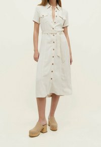 Robe chemise beige clair à manches courtes, avec un devant à boutons, deux poches poitrine et une ceinture en tissu. Associée à des sabots avec un talon en bois.