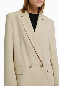 Beige dubbelgeknoopte blazer met een gladde textuur, scherpe revers en twee voorzakken. Voorzien van metalen knopen en een getailleerde pasvorm.