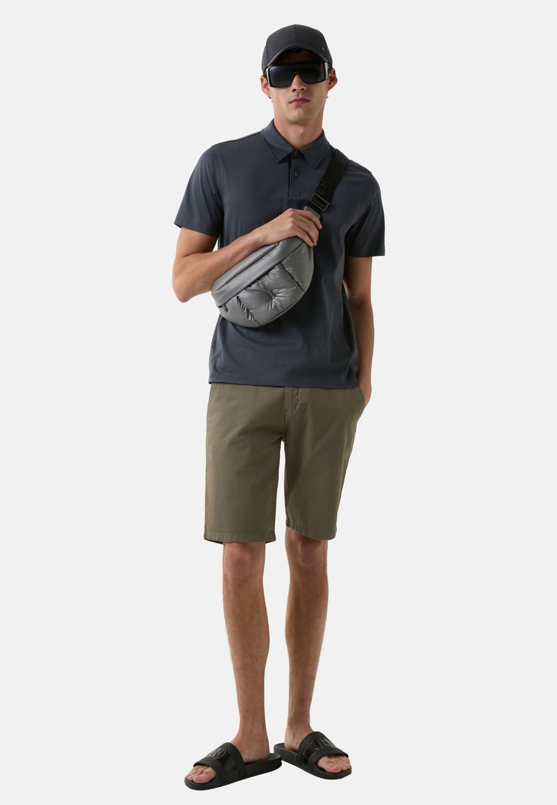 Grauer Puffer-Crossbody-Rucksack, marineblaues Polo-Shirt, olivgrüne Shorts und schwarze Sandalen. Dazu gehören eine schwarze Kappe und große Sonnenbrillen.