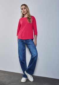 Czerwona bluzka z długim rękawem i okrągłym dekoltem, w zestawieniu z szerokimi niebieskimi dżinsami i białymi trampkami, na neutralnym tle.