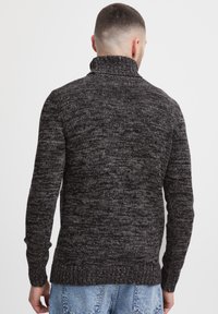 Grå strikket genser med høy krage, som har et m�ønster med tekstur og ribbestrikkede mansjetter, vist fra baksiden. Kombinert med denimjeans.