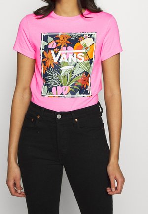 Osoba nosząca jasnoróżowy t-shirt Vans z kolorowym kwiatowym wzorem i czarne dżinsy z wysokim stanem, pokazana od szyi do bioder.