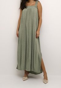 Robe longue verte en tissu texturé, décolleté carré et bretelles à volants. Comprend une fente sur le côté et tombe à la longueur des chevilles.