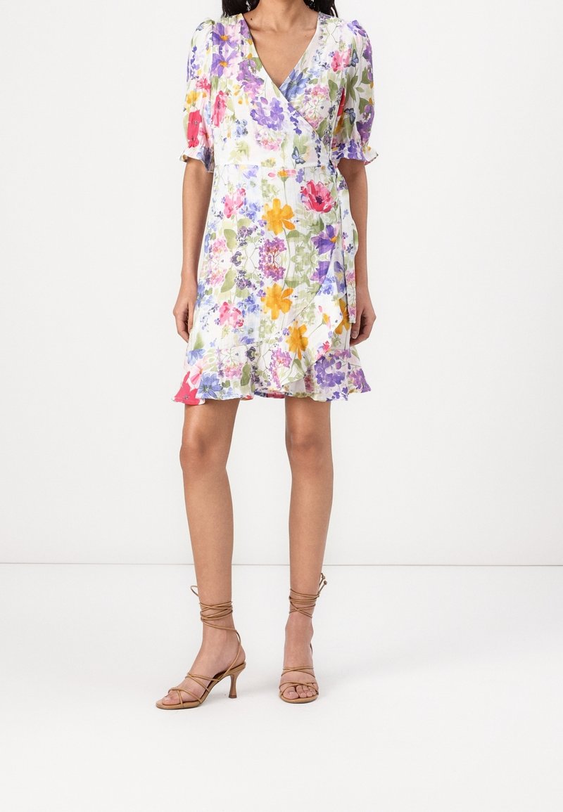 Femme vêtue d'une robe portefeuille blanche à imprimé floral coloré, manches bouffantes, ourlet à volants, et sandales à talons beiges à lanières.