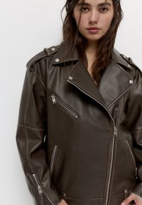 Junge Frau mit langen dunklen Haaren, die eine übergroße dunkelbraune Lederjacke mit silbernen Reißverschlüssen und Knöpfen trägt.