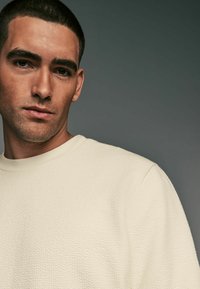 Lys beige tekstureret sweatshirt med ribbet halsudskæring, med et geometrisk mønster og en afslappet pasform, delvist vist på en model.