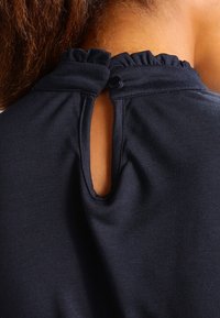 Mörk marinblått tyg med rund halsringning som har en volangdetalj och en nyckelhålsöppning bak, säkrad med en knapp.