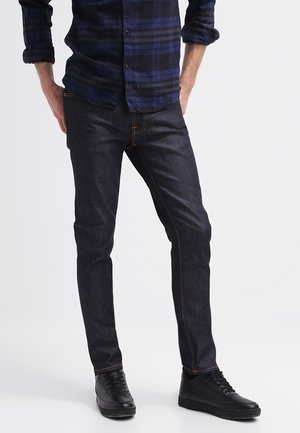 Mand iført mørkeblå jeans, sorte strukturerede sneakers og en blå og sort ternet langærmet skjorte med opbukte ærmer.