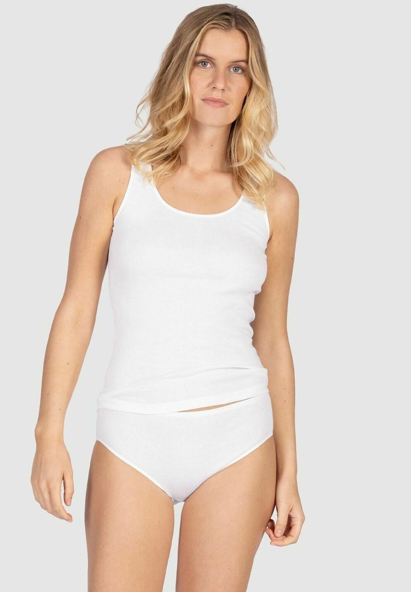 Blonde Frau, die ein weißes ärmelloses Top und passende weiße Unterwäsche trägt, steht vor einem schlichten hellgrauen Hintergrund.