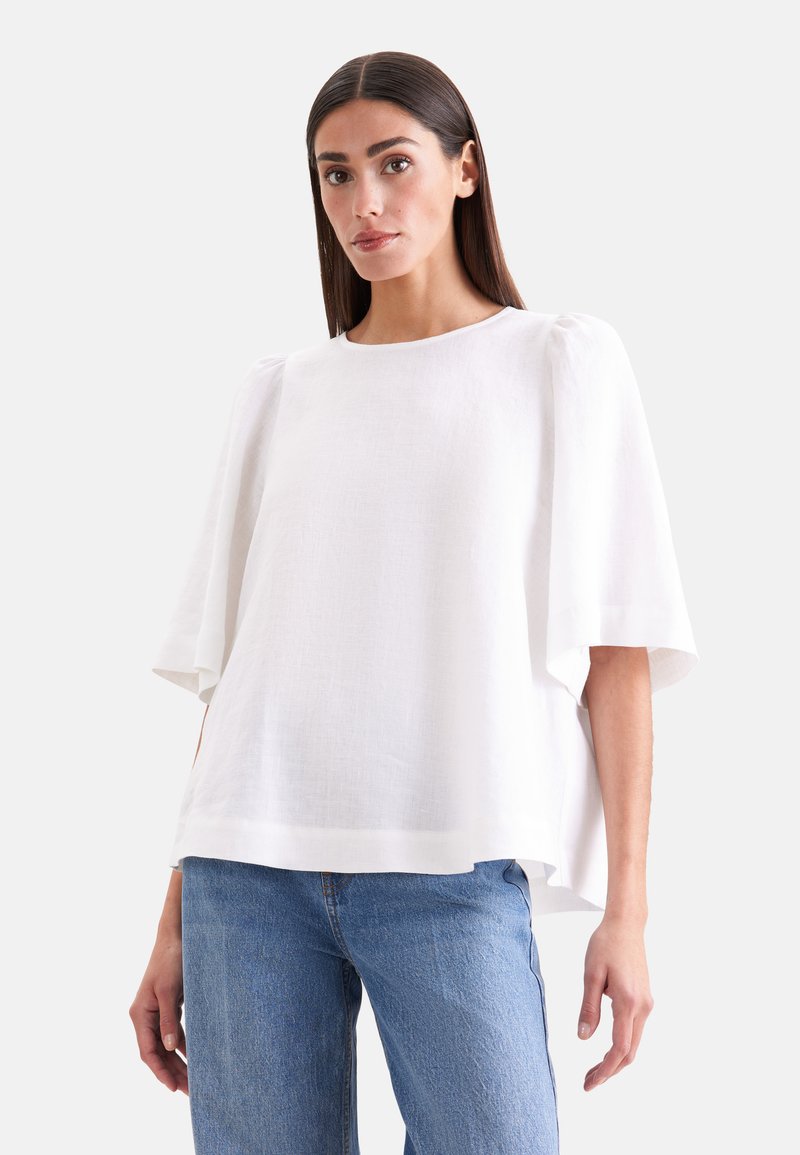 Femme portant un chemisier blanc ample à manches larges et un jean bleu, debout devant un fond clair uni.