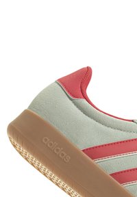 Adidas-sneaker med en mjuk grön mockaöverdel, röda läderdetaljer och en texturerad gummisula. Har tre vita ränder på sidan.
