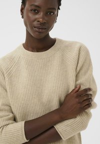 Pull en tricot côtelé beige avec un col rond et des manches raglan, présentant une texture douce et une coupe ample, porté avec les bras croisés.