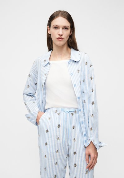 Woman wearing light blue and white striped pajama set with embroidered bees, white undershirt, standing with one hand in pocket.