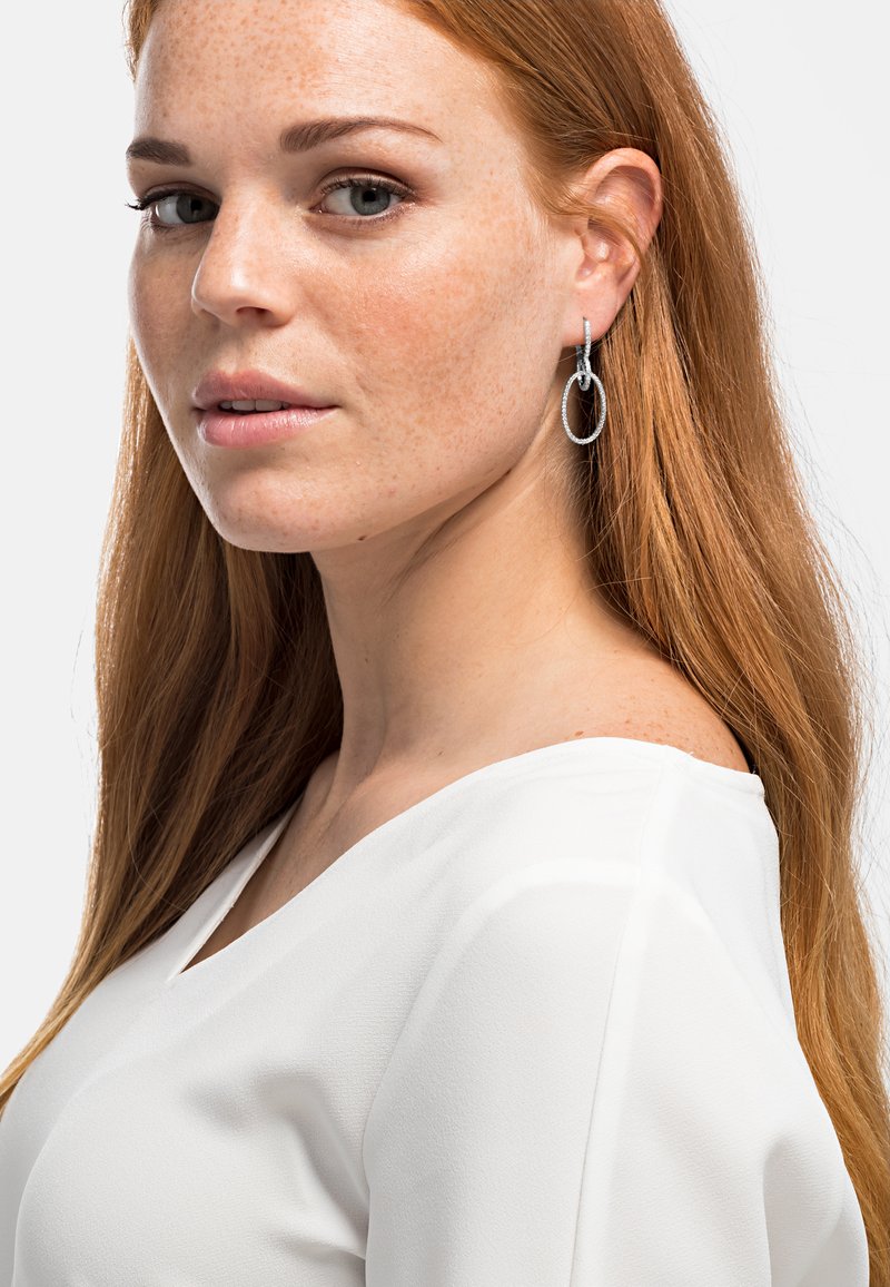 Silberne Creolen mit strukturierter Oberfläche, getragen von einer Person mit langen, glatten roten Haaren und einer weißen Bluse mit V-Ausschnitt.