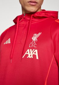 Červená mikina s kapucí z hladké látky s logem Liverpool FC a nápisem „AXA“ v krémové barvě. Obsahuje zip u límce a jasně oranžové detaily.