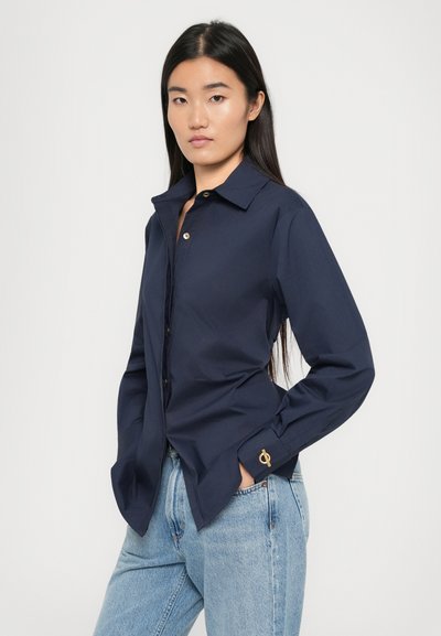 Camicia blu navy con colletto classico, maniche lunghe, leggero lucido e un accento dorato sul polsino. Indossata con jeans azzurri chiari.