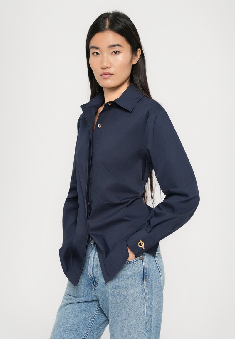 Chemise bleu marine à boutons avec un col classique, manches longues, léger éclat et une touche dorée sur le poignet. Portée avec un jean bleu clair.