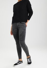 Suéter negro de punto trenzado, jeans skinny grises con acentos en canalé y zapatillas negras y blancas; atuendo casual moderno con texturas variadas.