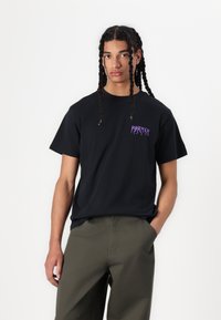 Zwarte katoenen T-shirt met een paarse grafische tekstlogo op de borst. Het model combineert het met olijfgroene broeken, wat een relaxte pasvorm laat zien.